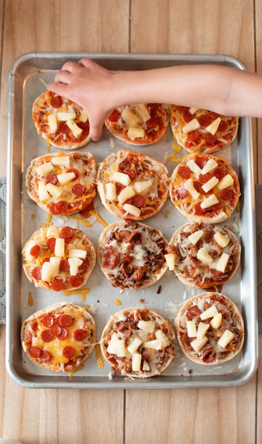 A child's hand reaching out for an English muffin pizza on baking sheet.