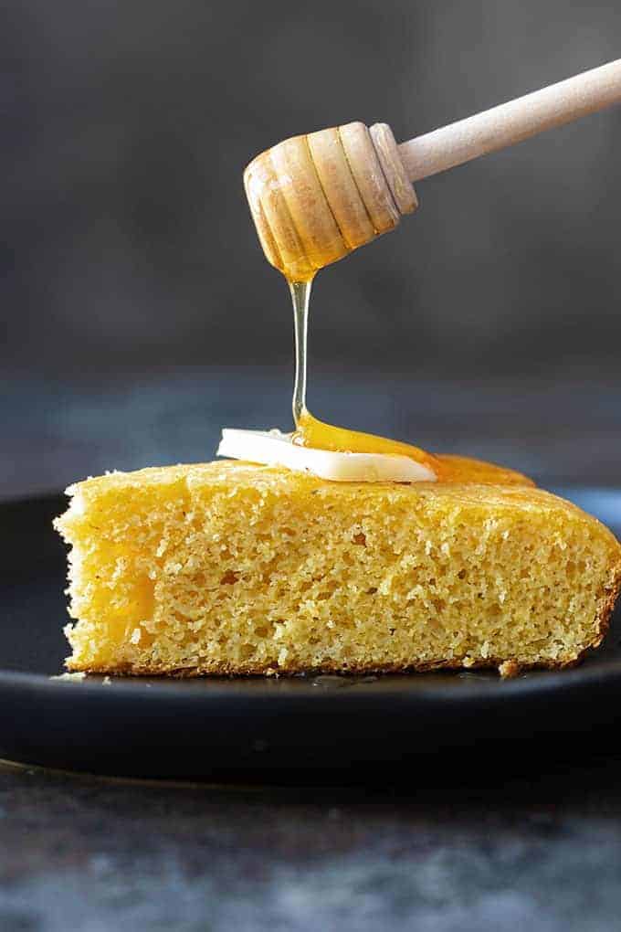 A slice of cornbread on a black plate being drizzled with honey.