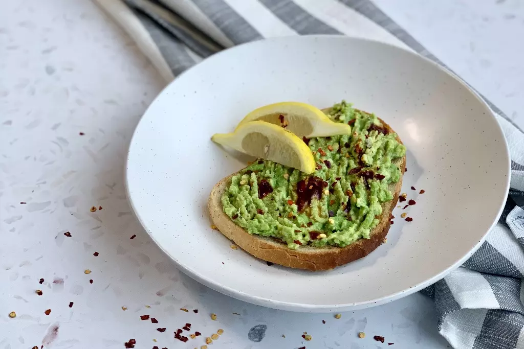 Avocado on toast with two slices of lemon on a white plate.