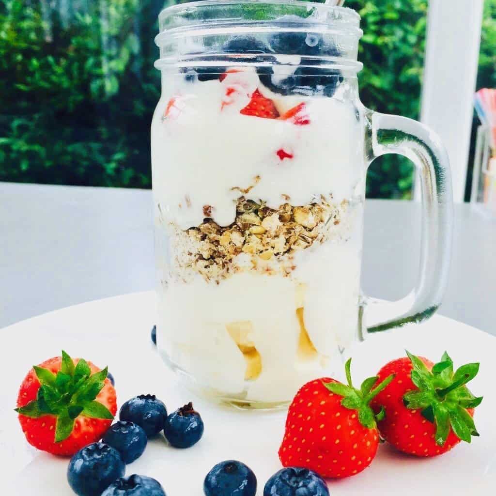 Cereal sundae topped with fresh fruits in a mason jar next to strawberries and blueberries.
