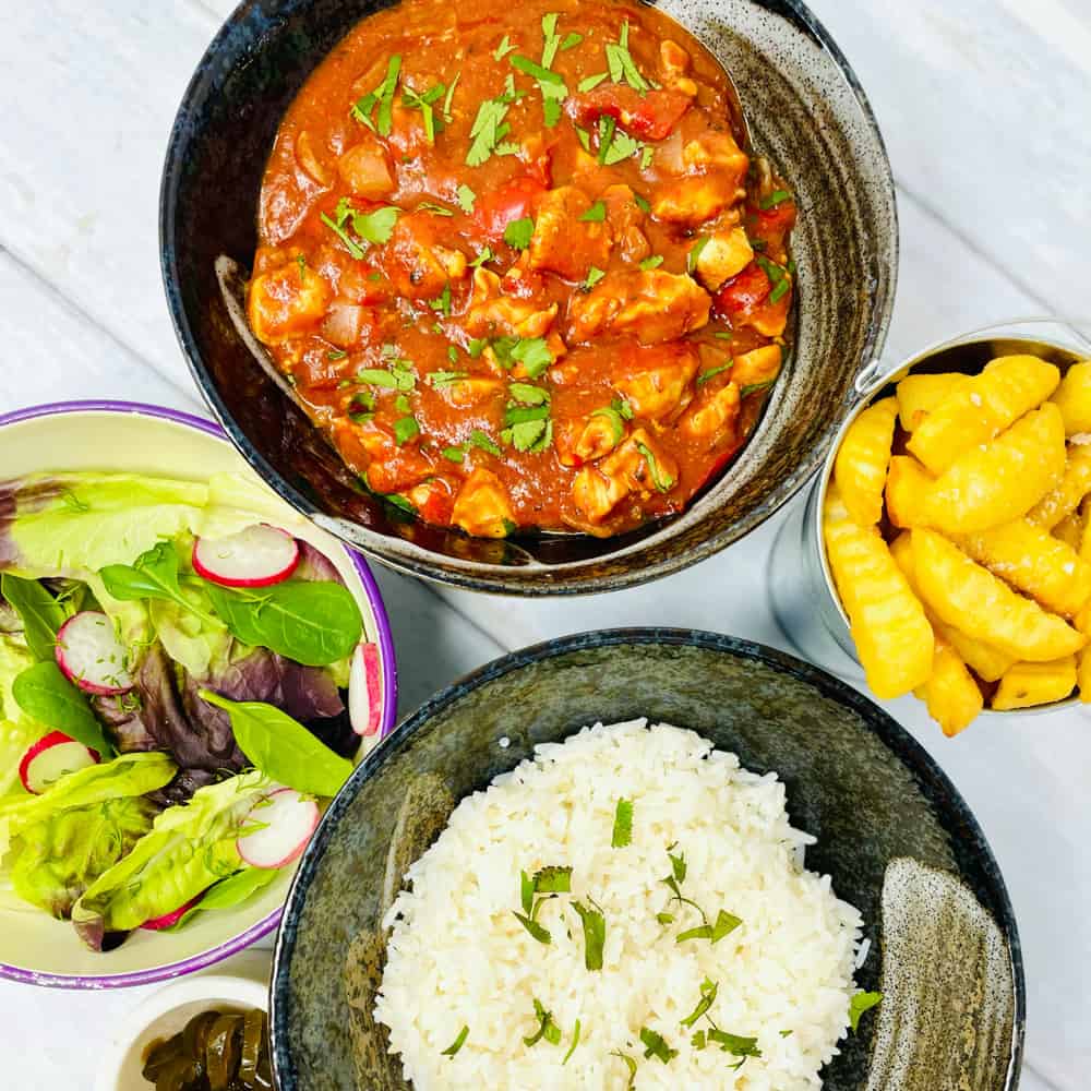 A bowl of green salad, a plate of rice, potato wedges and a plate of canjun chicken.