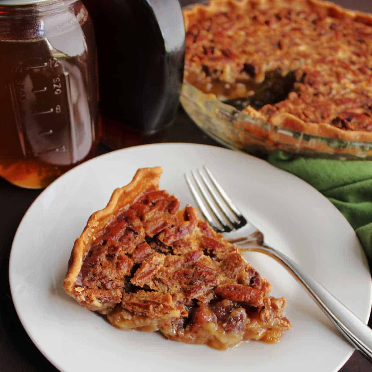 A slice of pecan pie on a plate with a fork, a whole slice of pecan pie in the background.