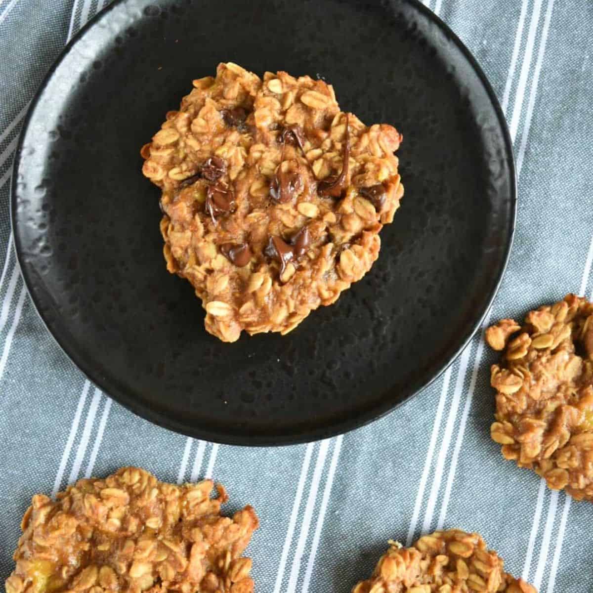 A piece of 3-ingredient peanut butter oatmeal cookies in a black plate surrounded by three pieces of the same cookies on a white and blue striped cloth.
