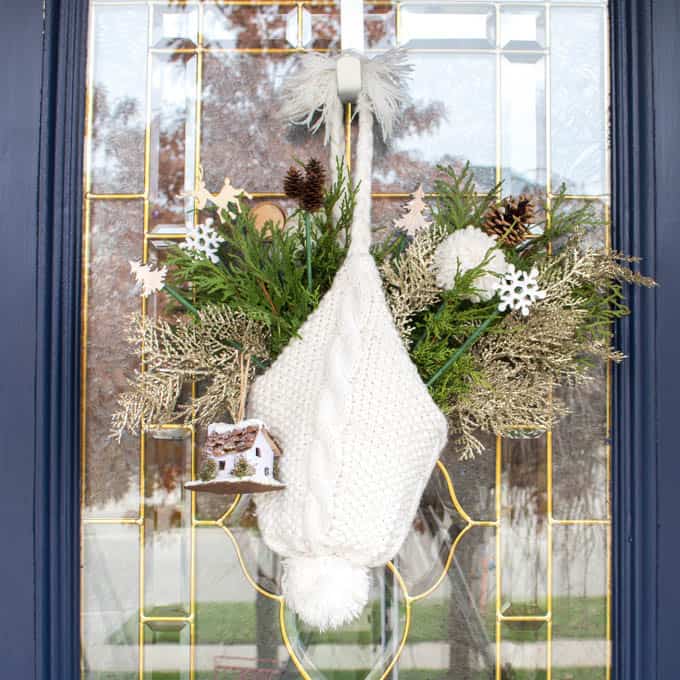 A white winter hat decorated with evergreens hanging upside down from a glass door.