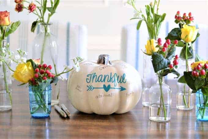 White pumpkin with "thankful" written on it in blue ink next to white and blue glass vases with yellow roses and red flowers.