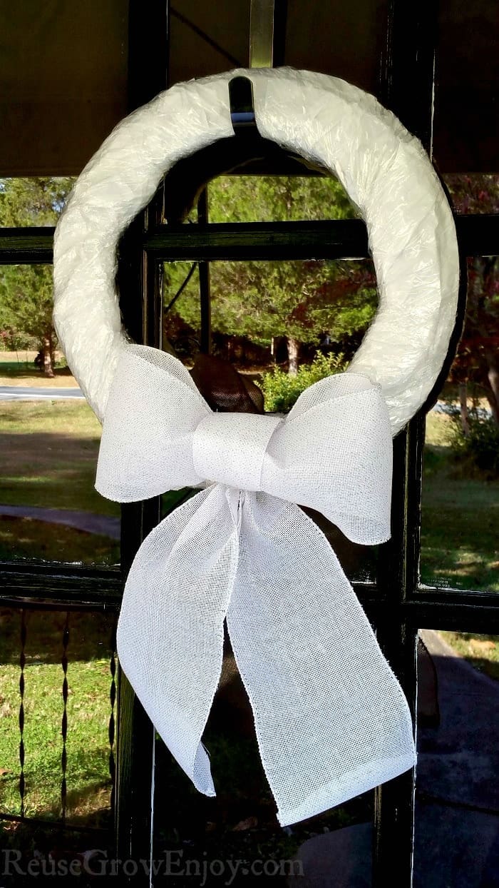 White wreath with large white bow hanging from a glass French door that reflects the images of the surrounding trees.