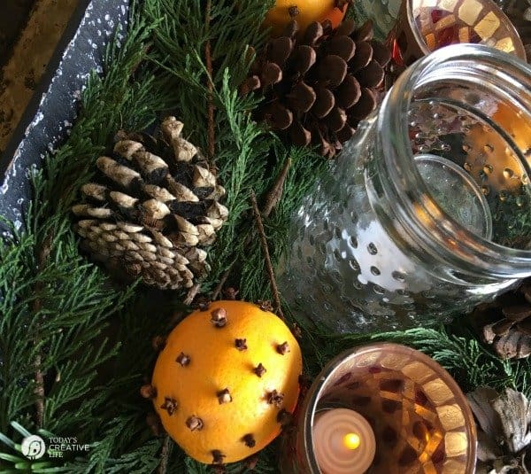 A clear glass in the middle of pinecones, oranges pinned with cloves and small branches of evergreen plant.