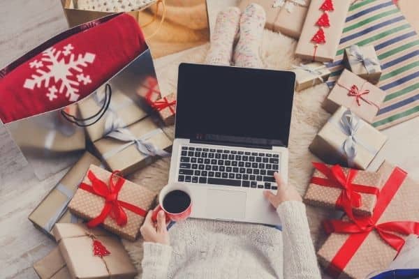 A person holding a laptop on one hand and a cup of coffee on the other hand surrounded by gift boxes with red and silver ribbons.