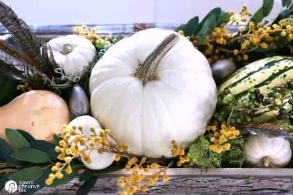 Large white pumpkin in the middle of small white pumpkins and different varieties of squash designed with leaves and yellow flowers in a wooden tray.