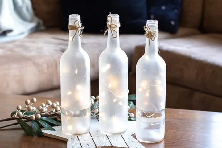 Fairy lights inside the three frosted wine bottles that are placed on top of the wooden table next to a branch of golden berries decoration.