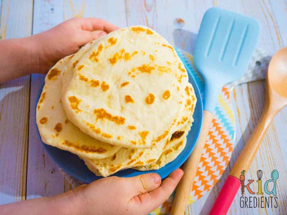 A person holding a blue plate with stack of flatbread next to a blue and a wooden spatula.