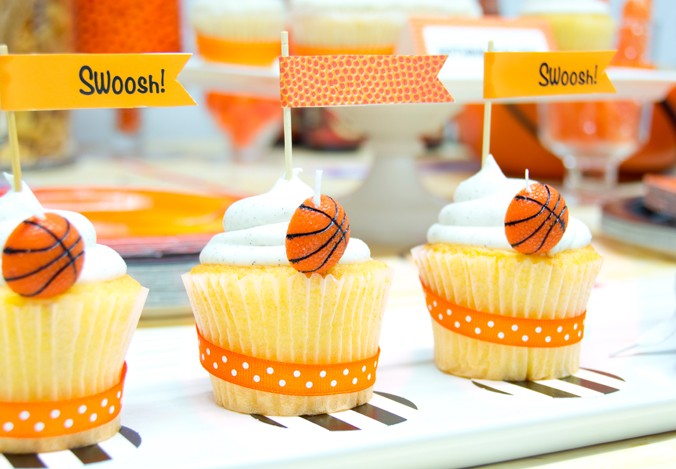 Three basketball cupcakes with two of them having a small flag that says swoosh.