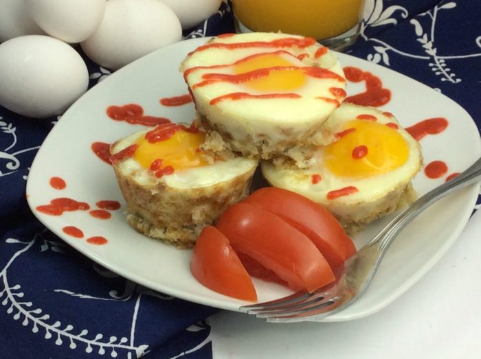 Three egg muffins on a white plate with tomato slices on the side and eggs as background.