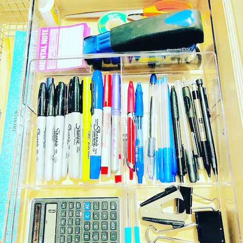 A clear desk drawer organizer for pens, sharpie, binder clips, calculator, pair of scissor, tape notepad and stapler.