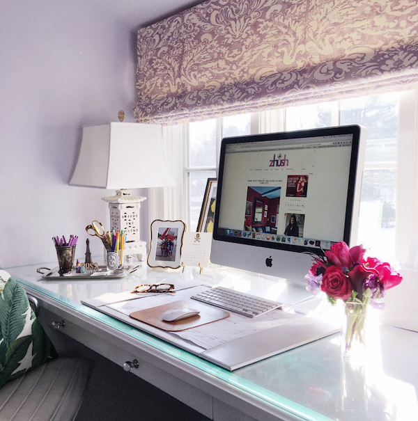 A clean office desk with an iMac, pink flowers on one side and a white lamp, photo frame and pen holders on the other side.