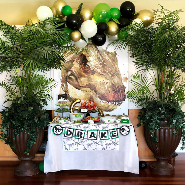Dinosaur themed birthday party table with an image of a T-rex as background and real-life plants in pots on both sides.