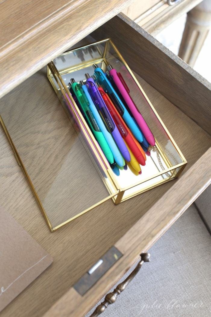 A clear pen organizer with colored pens inside an opened wooden desk drawer.