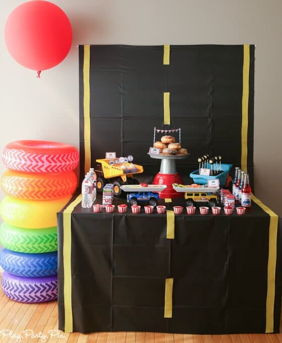 Road -themed birthday party with a snack table that has construction trucks filled with cupcakes and cakepops and a plate of donuts in the middle next to a colorful stack of plastic car wheels.
