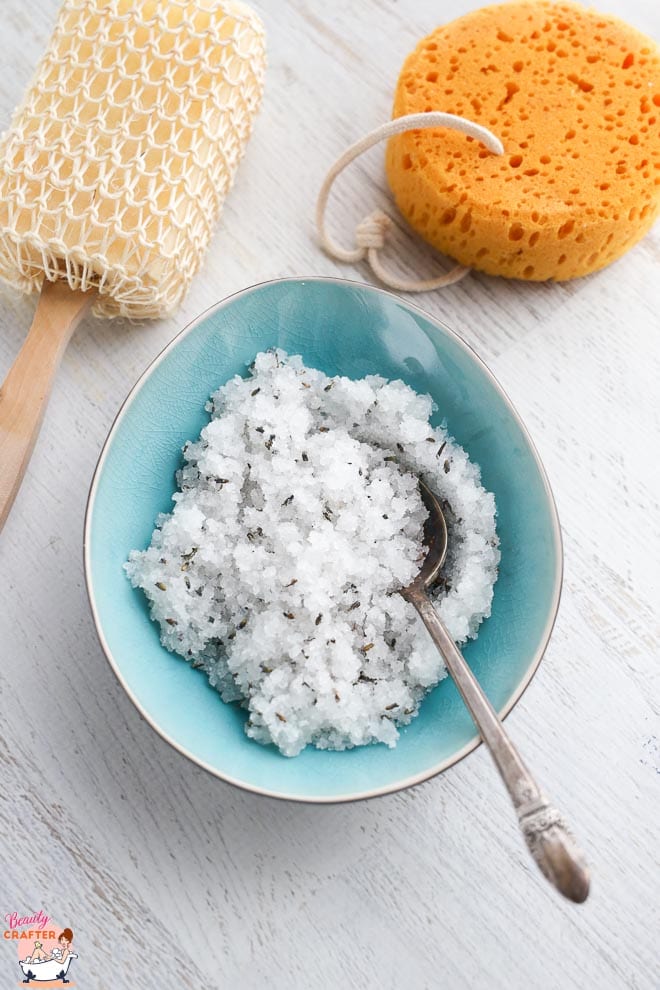 Sea salt scrub in a blue bowl with spoon next to a loofah and sponge bath.