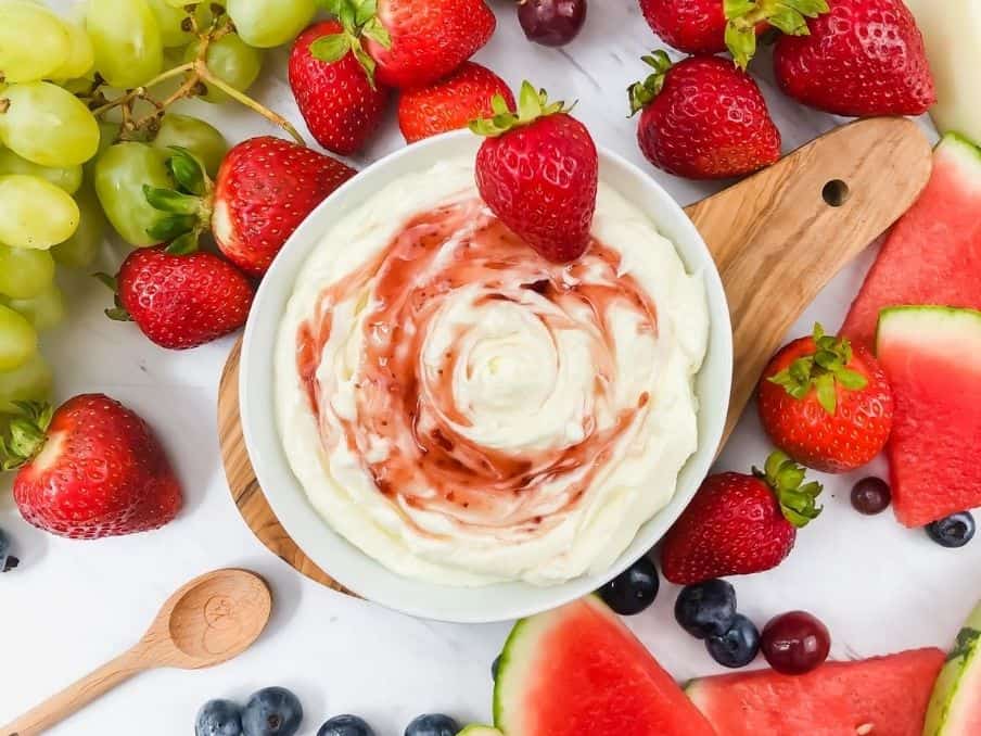 Fruit dip swirl in a white bowl with strawberries, blueberries, green grapes and watermelon slices on the side.