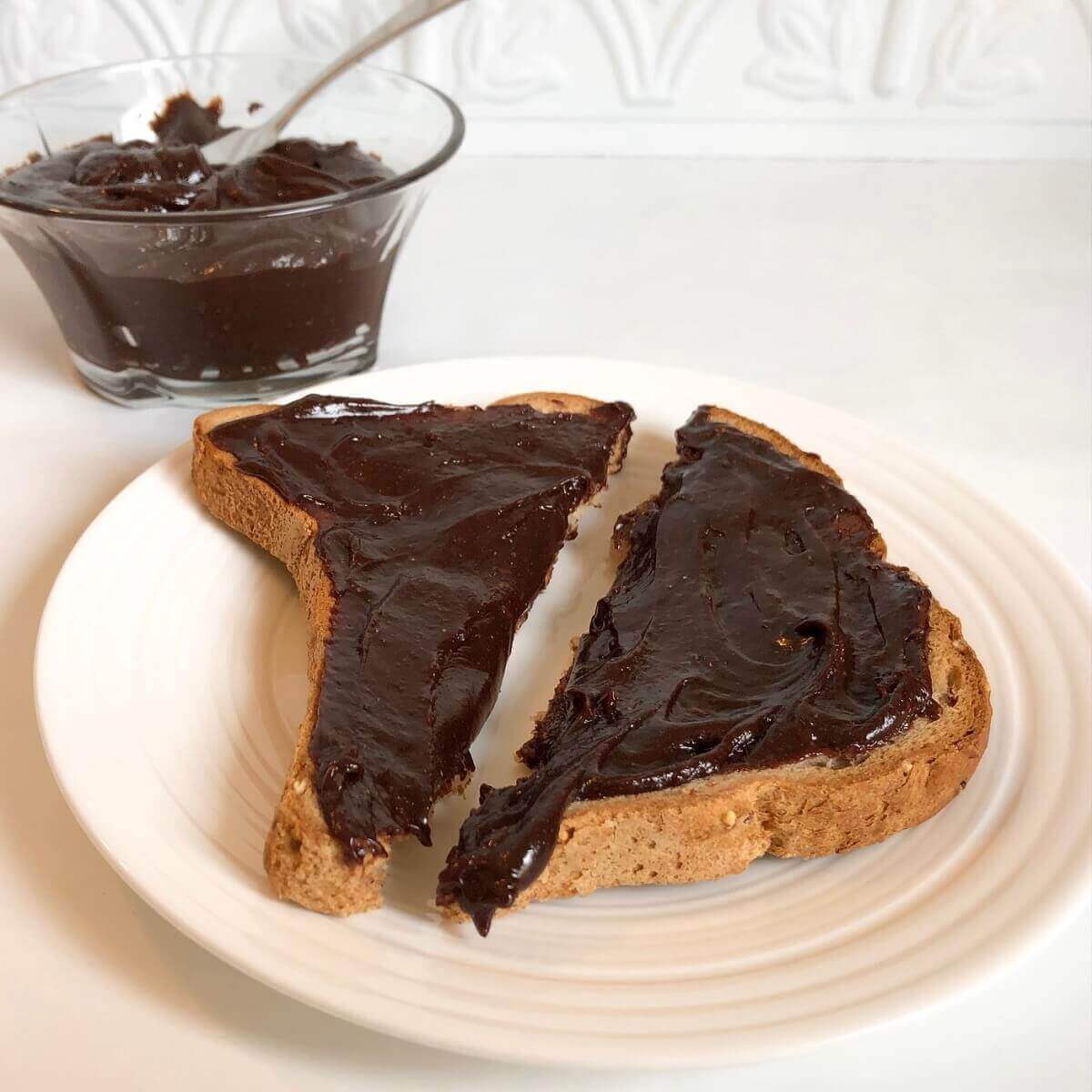 Toast with chocolate spread sliced in half on a white plate next to a bowl of chocolate spared.