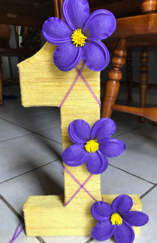 Number one table decor wrapped with yellow yarn and decorated with three purple flowers.