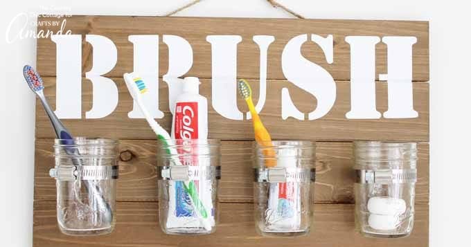Four mason jars on wooden plaque with word "BRUSH" for bathroom organization.