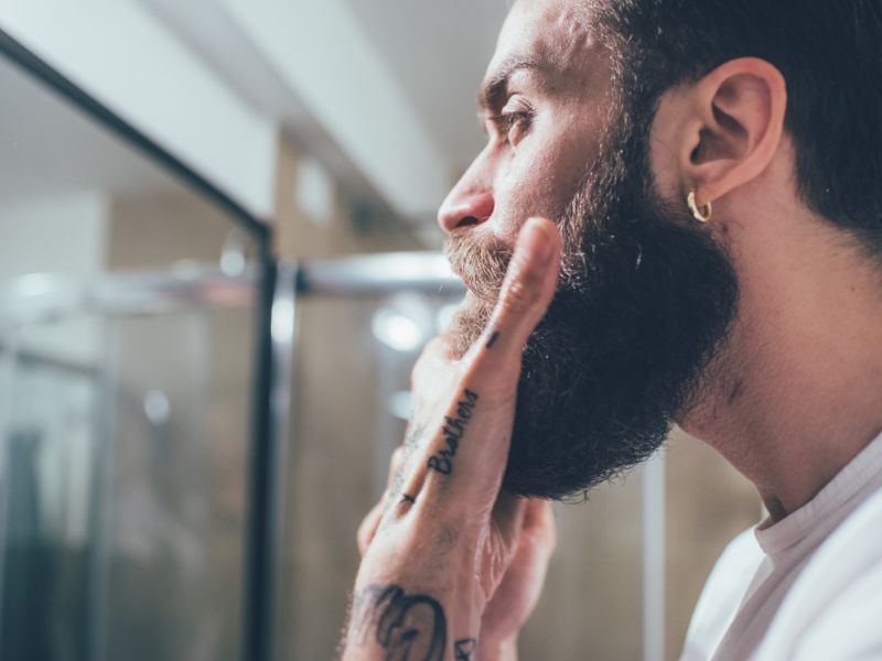 A man with a beard in front of a mirror holding his chin.