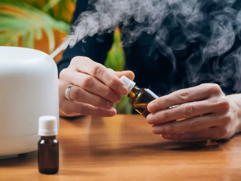 A person holding a bottle of clean cotton essential oil. White diffuser misting next to a small brown bottle of essential oil on wooden table.