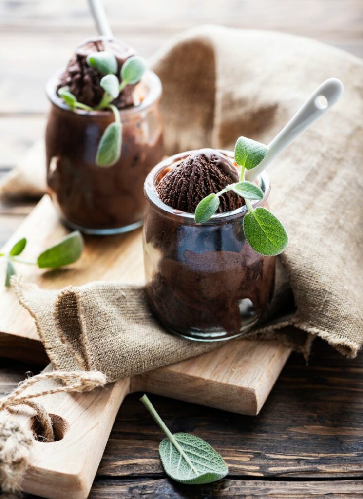 Two jars of chocolate sage ice cream with spoon and green herb garnish on a cutting board with burlap design.