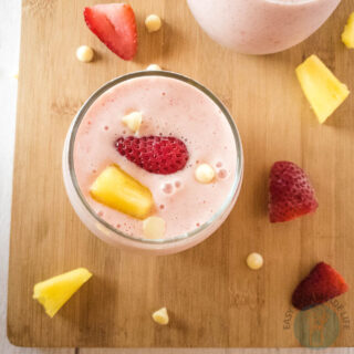 Two glasses of bahama mama smoothie on a wooden chopping board surrounded by slices of pineapples and strawberries.