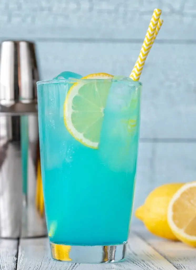 A glass of electric blue lemonade drink with yellow straws and lemon wedges. Cocktail shaker and lemons in the background.