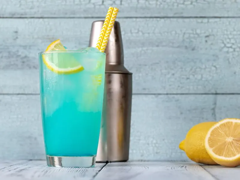 A glass of electric blue lemonade drink with yellow straws and lemon wedges. Cocktail shaker and lemons in the background.