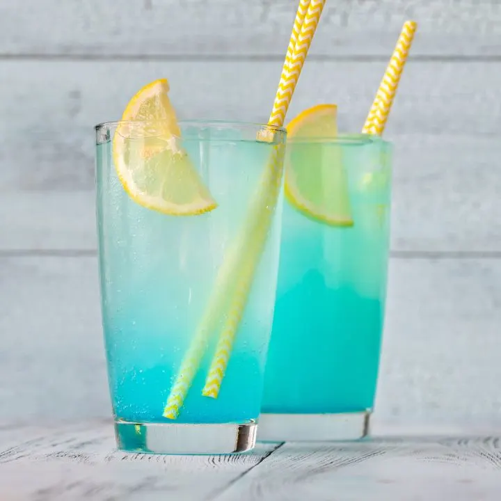Two glasses of electric lemonade drink with yellow straws and lemon wedges on white-painted wooden table.