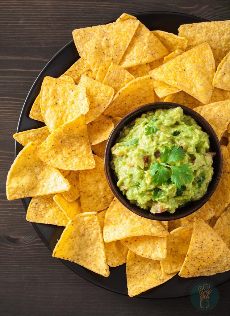 A bowl of guacamole in the middle of chips in a black plate.