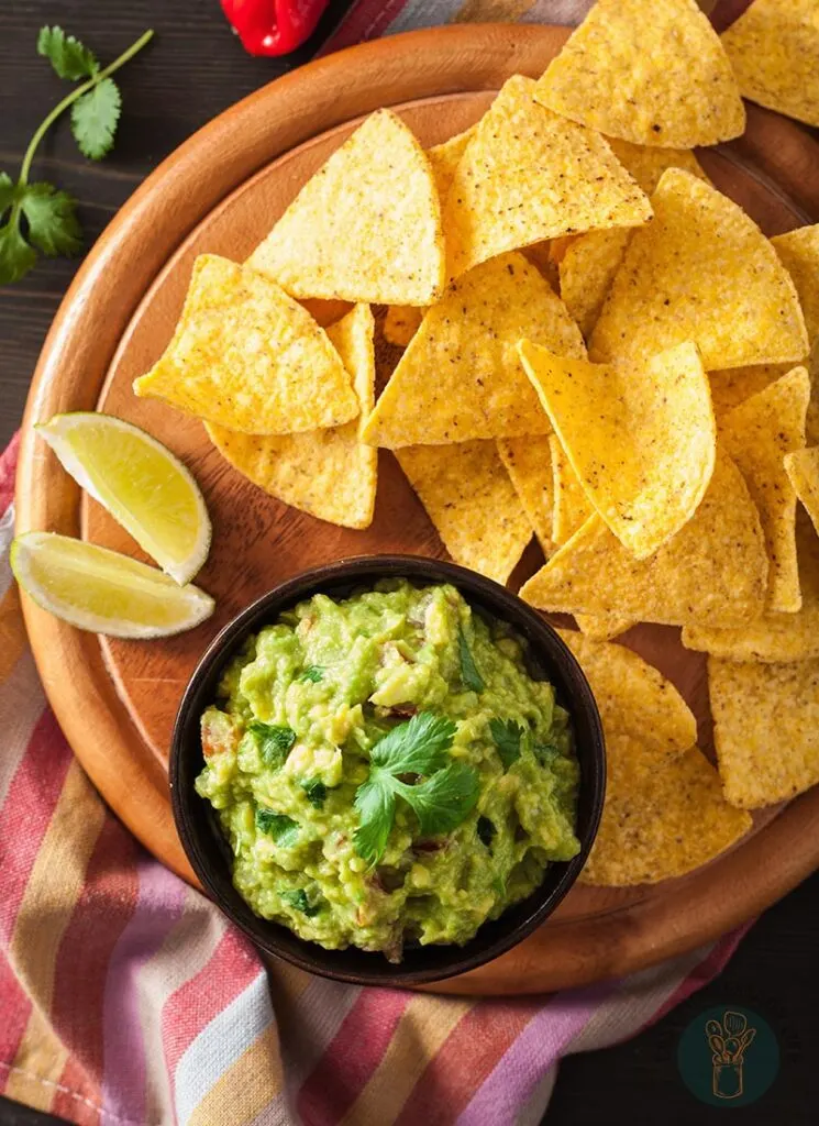 A bowl of guacamole next to chips and two lemon slices in a wooden plate.