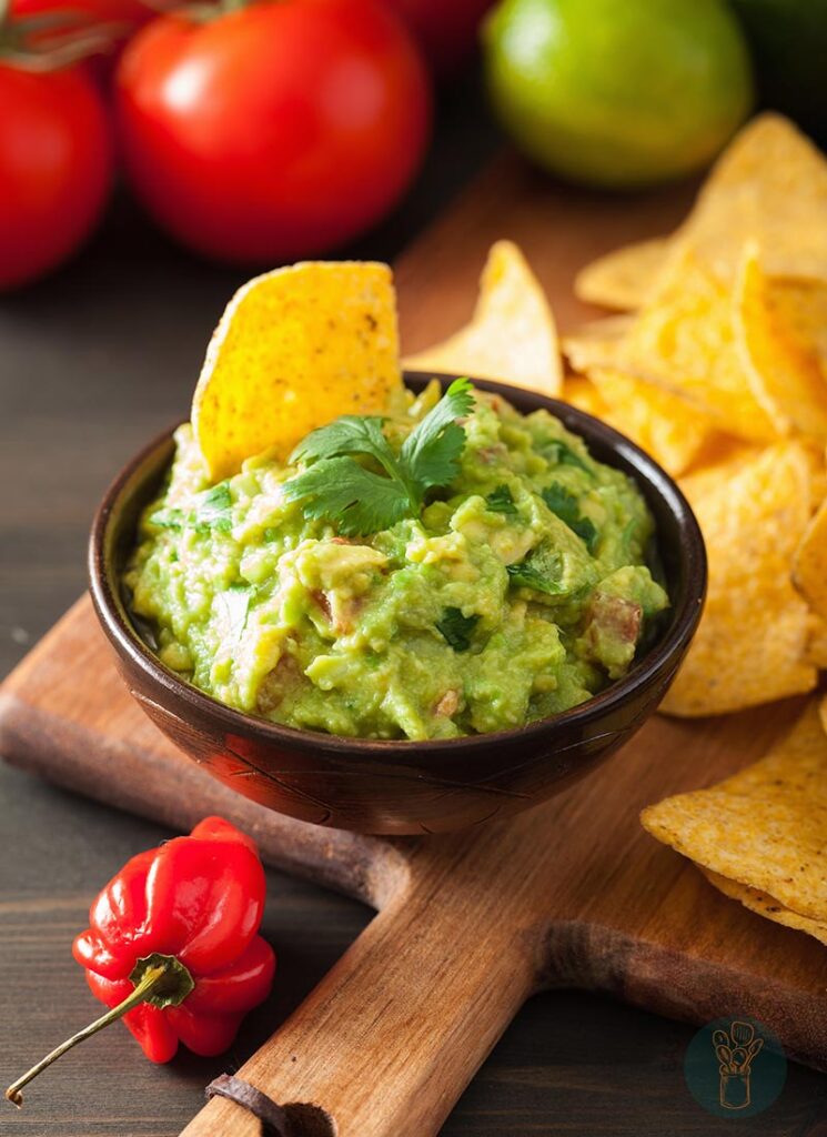 A bowl of guacamole with cilantro garnish and a piece of chip dipped in it on a wooden chopping board with chips.
