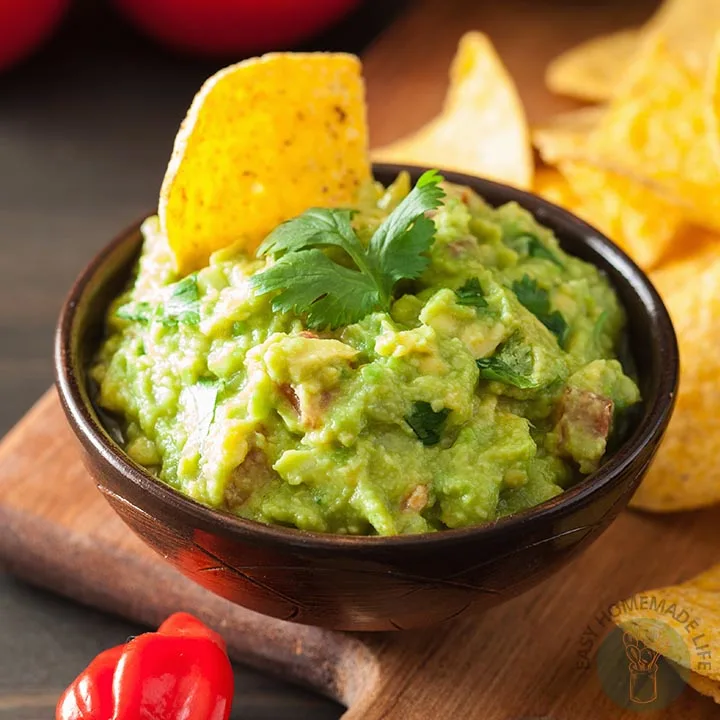 A bowl of guacamole with cilantro garnish and a piece of chip dipped in it on a wooden chopping board.