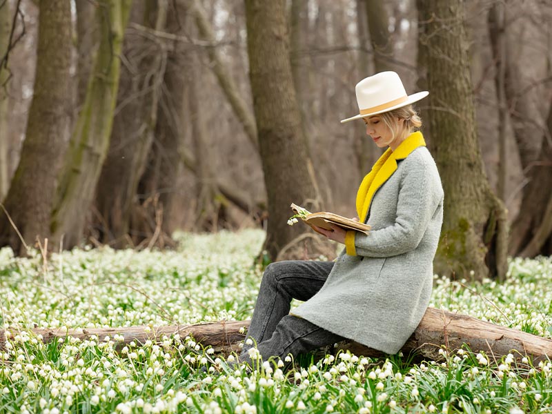 A woman wearing a hat and a grey coat sitting on a branch of tree and reading a book in the meadow.