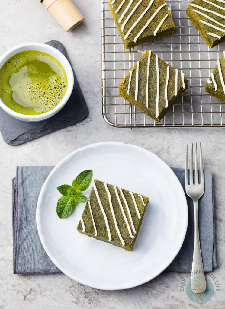 A square slice of matcha brownie garnished with mint in a white plate next to a fork, a cup of matcha tea, and slices of matcha brownies in a cooling rack.