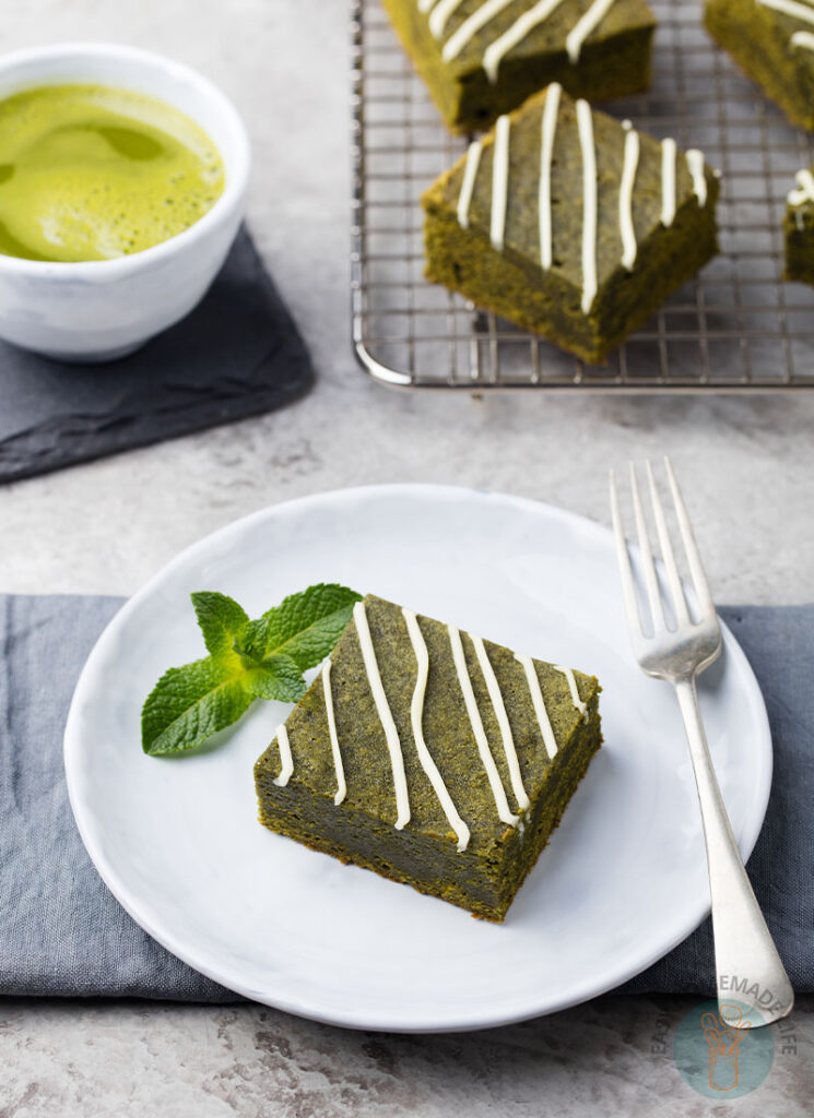 A square slice of matcha brownie garnished with mint in a white plate next to a fork, a cup of matcha tea, and slices of matcha brownies.