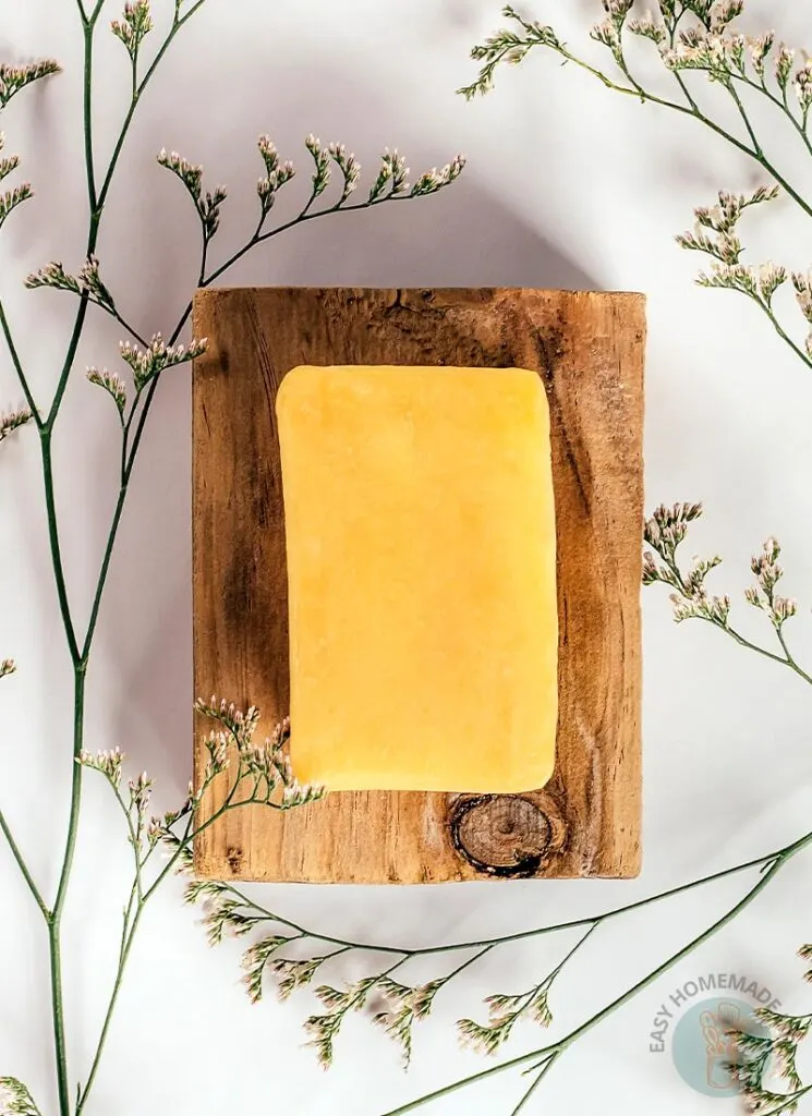 A bar of turmeric soap on a wooden board surrounded by green stems of plants with tiny flowers.