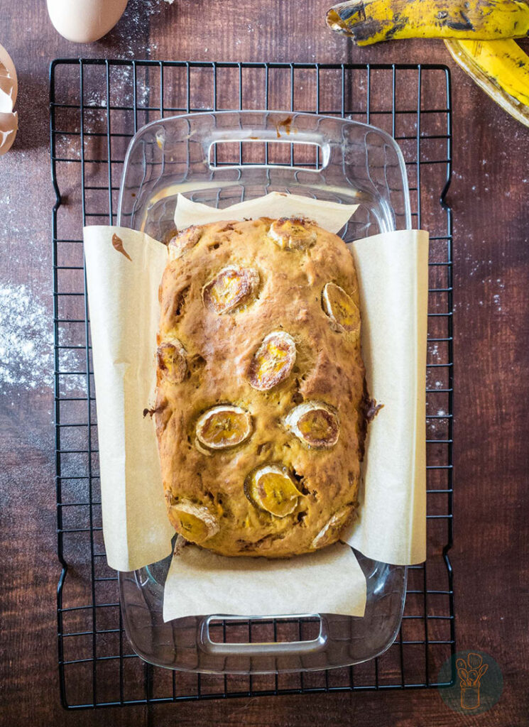 A whole banana bread in a baking dish placed on a cooling rack with ripe banana background.