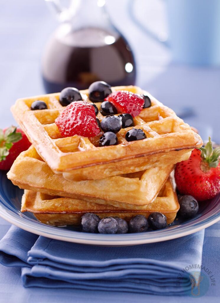 Three dairy-free waffles in a stack garnished with strawberries, blueberries and syrup in a blue plate next to a bottle of syrup.