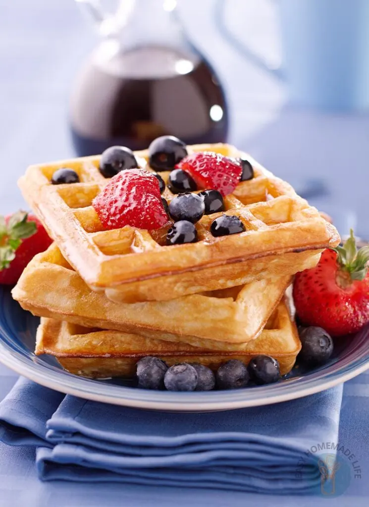 Three dairy-free waffles in a stack garnished with strawberries, blueberries and syrup in a blue plate next to a bottle of syrup.
