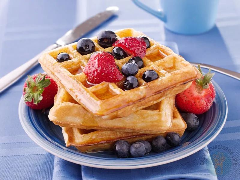 Three dairy-free waffles in a stack garnished with strawberries, blueberries and syrup in a blue plate.