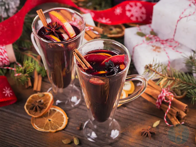 Two glasses of deep red drinks with slices of orange and cloves surrounded by holiday decorations.