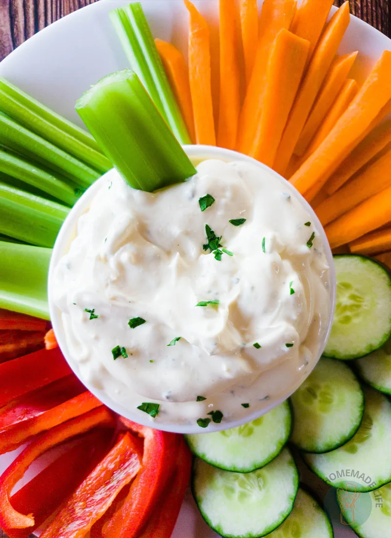 3-ingredient easy onion dip with a celery stick in it, in the middle of a plate full of celery sticks, carrots and red bell pepper cut in stripes and cucumbers cut in circle.