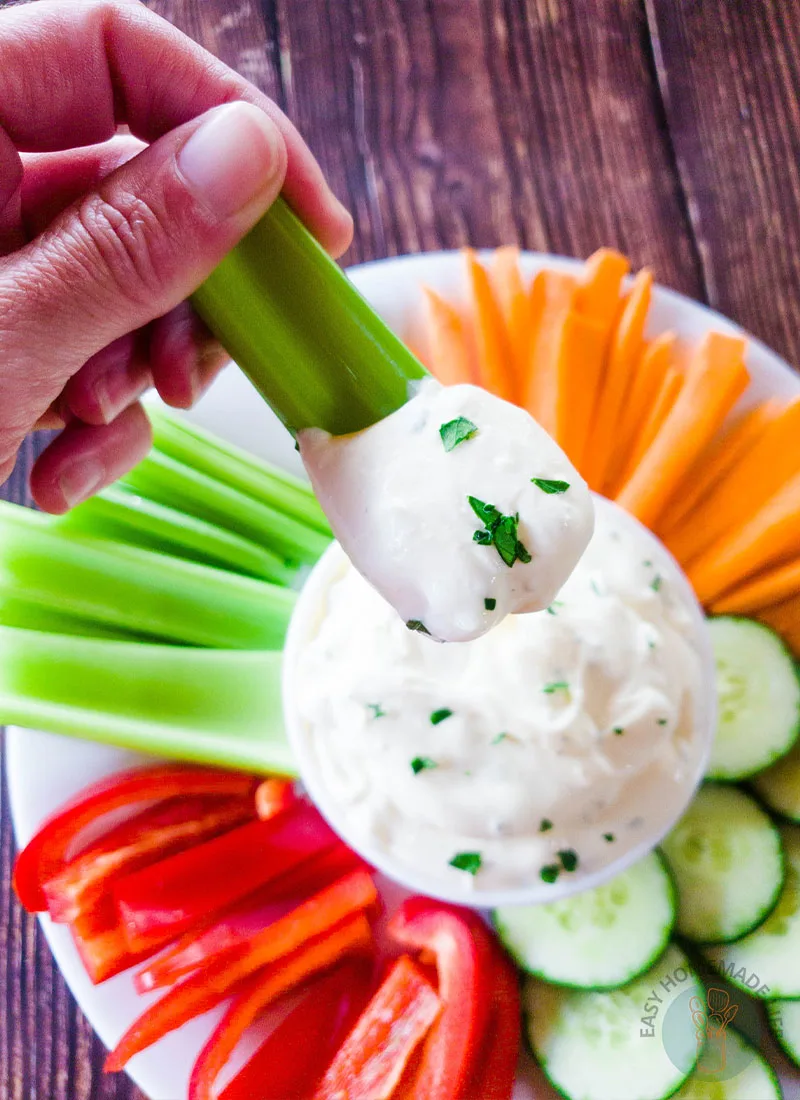 A hand holding a celery stick with an onion dip. A vegetable platter with 3-ingredient onion dip as the background.