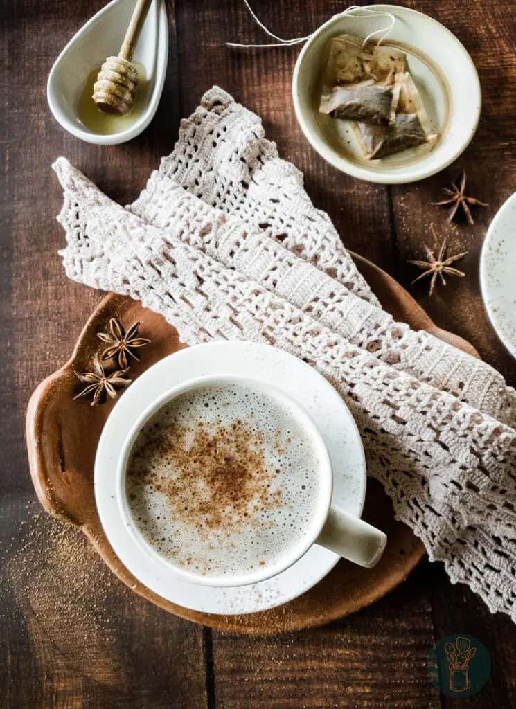 Hot chocolate chai tea in white teacup on clay dish with lace napkin next to tea bags and a honey dipper.